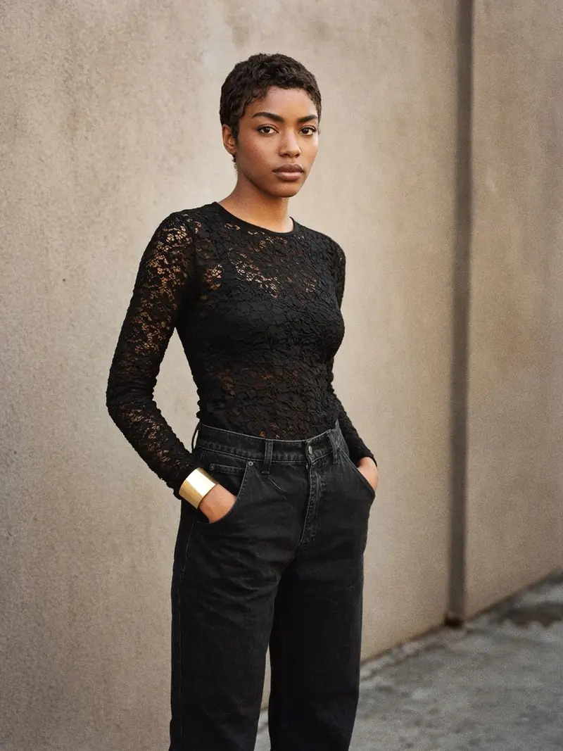 Illustration, mannequin fictif : Une femme en top noir en dentelle à manches longues doublé, jean droit noir taille haute et bottines noires, une large manchette en or jaune au poignet.