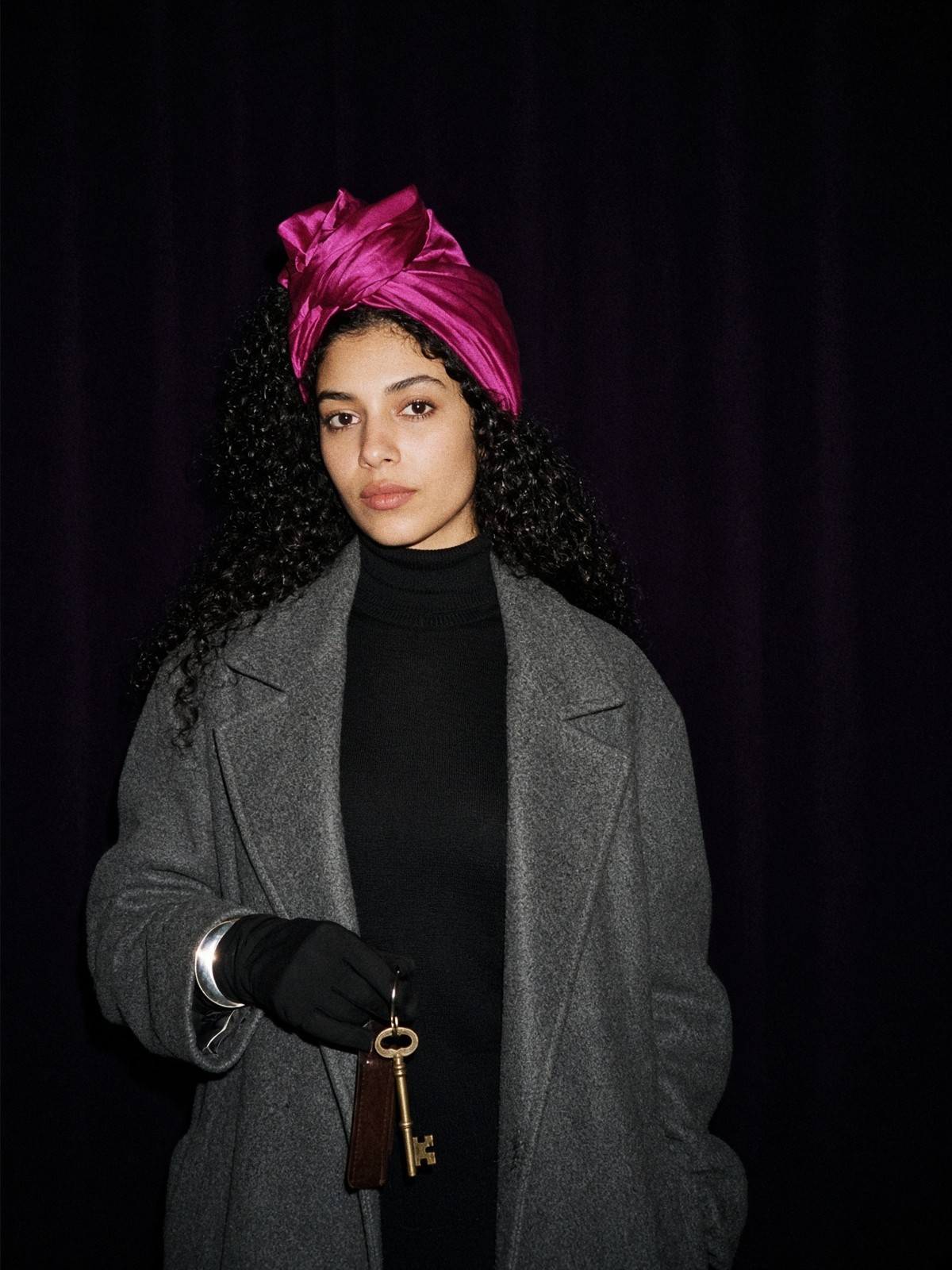 Mannequin en foulard de soie fuchsia noué haut en turban, col roulé noir, manteau gris et manchette argent