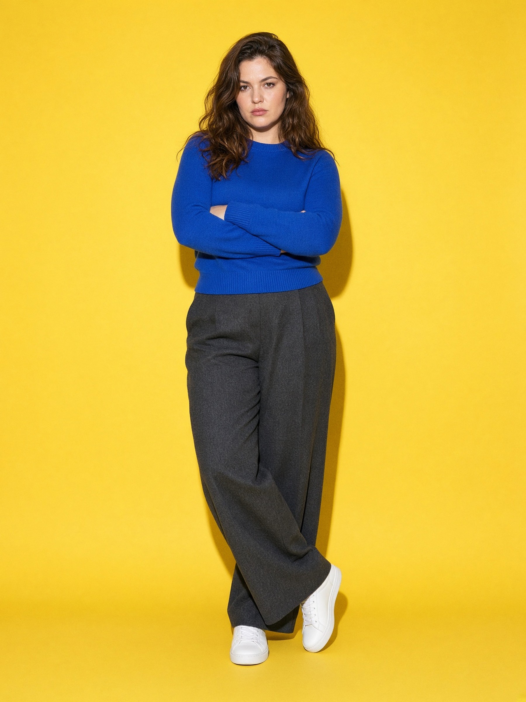 Mannequin en pantalon large anthracite, pull bleu cobalt et baskets blanches en cuir, un tabouret haut posé sur l'épaule, dans un studio nu