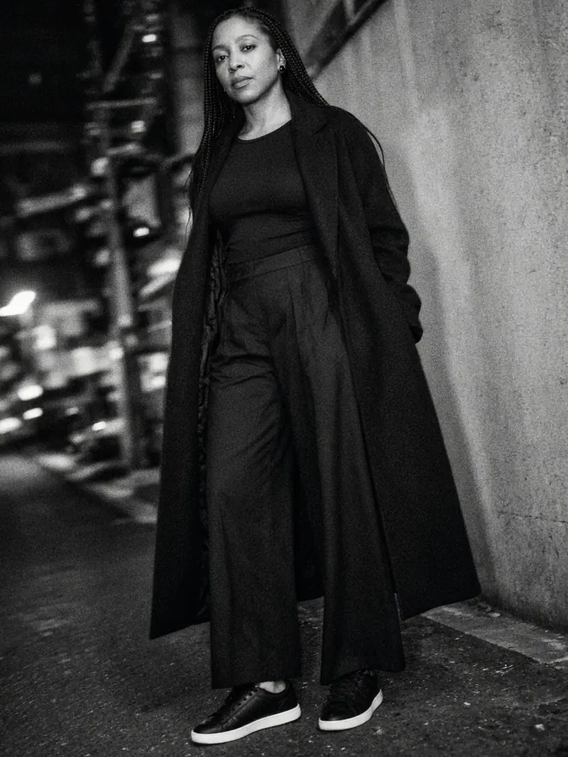 Illustration, mannequin fictif : Une femme en haut noir à manches longues, pantalon large en jersey noir, baskets noires et long manteau noir ouvert, debout dans une rue la nuit.