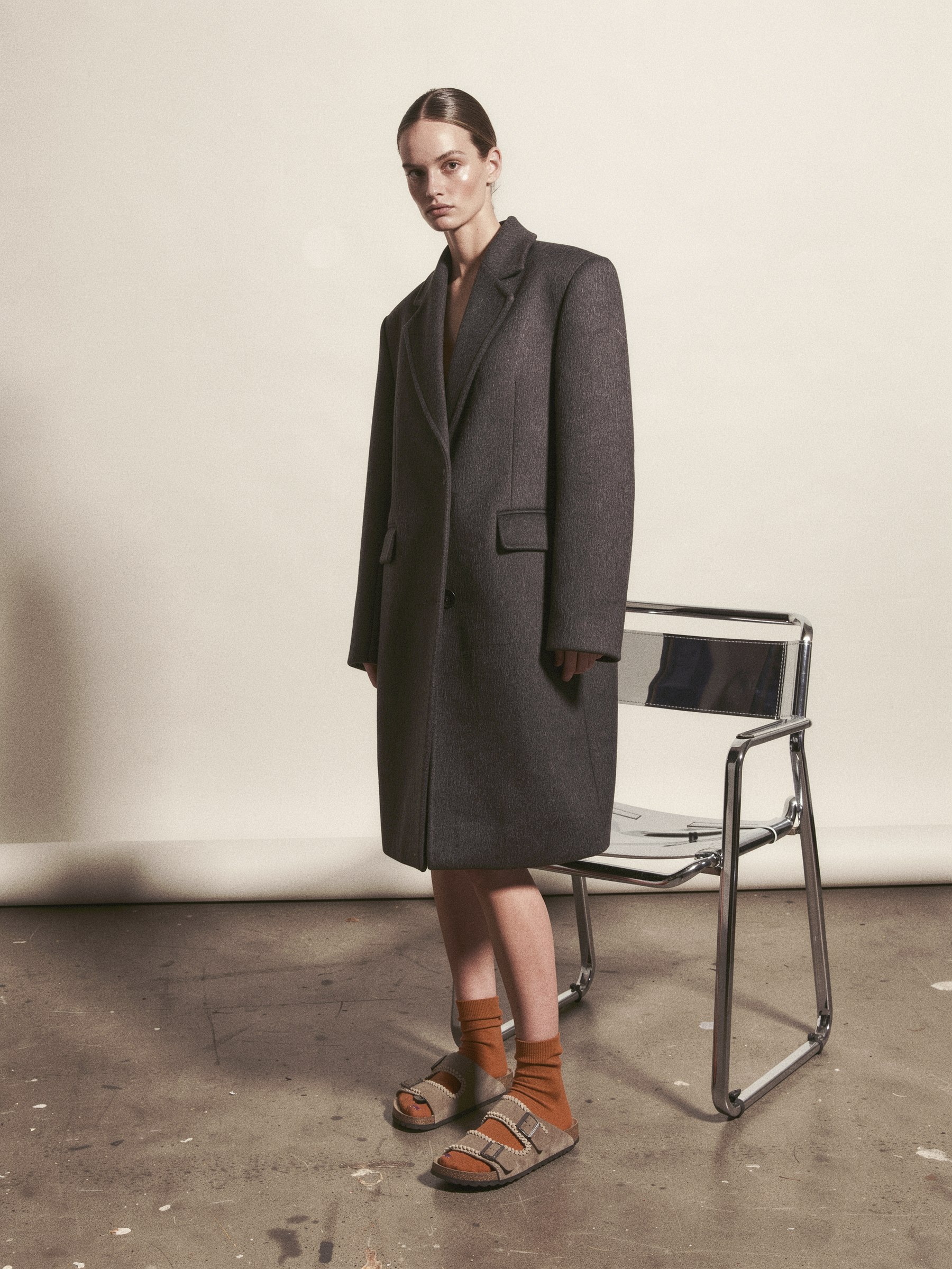 Mannequin en manteau structuré portant une sandale à franges en suède avec une chaussette fine, debout dans un décor de studio épuré.