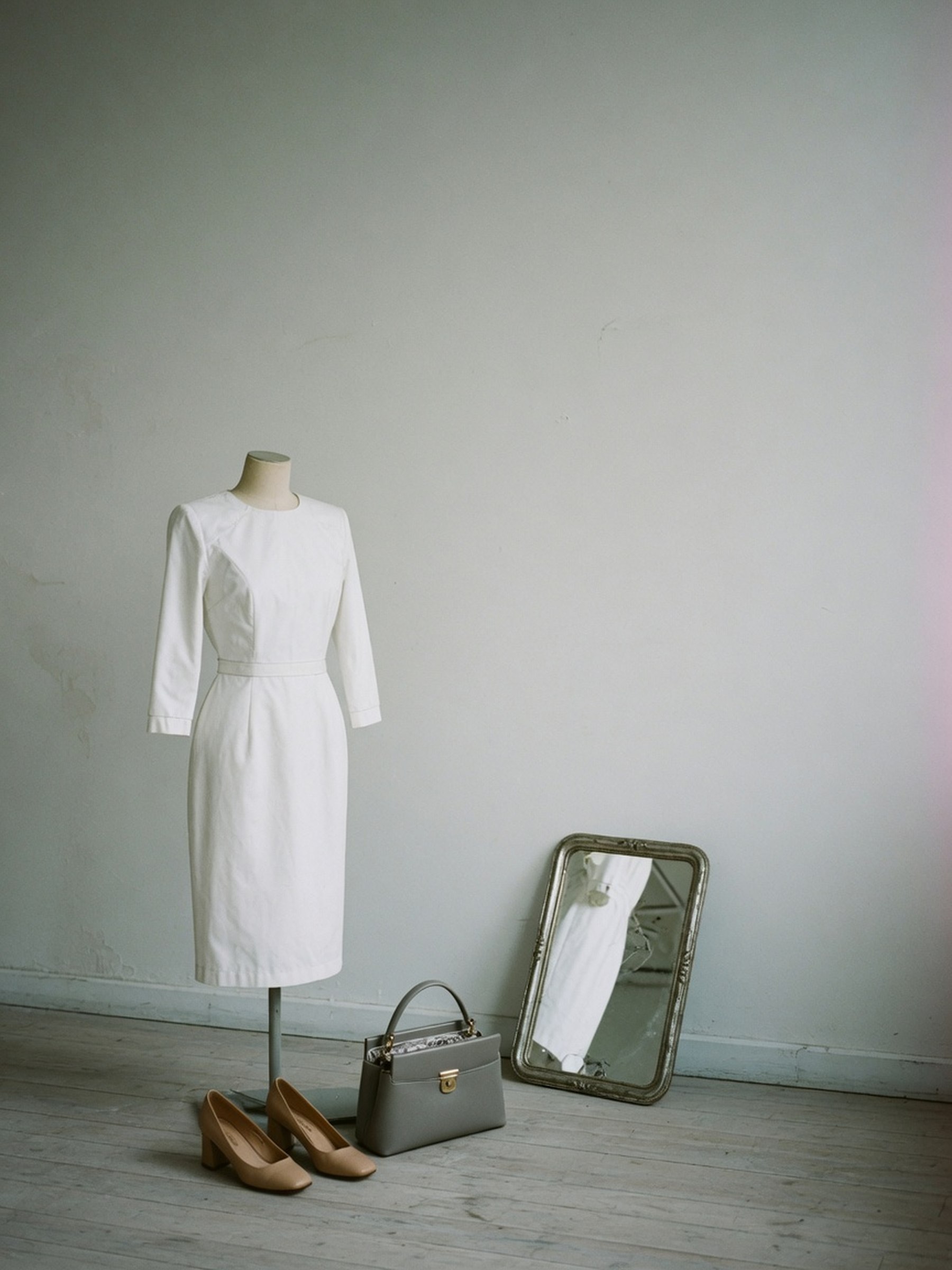 Nature morte éditoriale d'une robe blanche droite à manches trois-quarts posée sur un mannequin de couturière, avec escarpins beige et sac structuré gris au sol.
