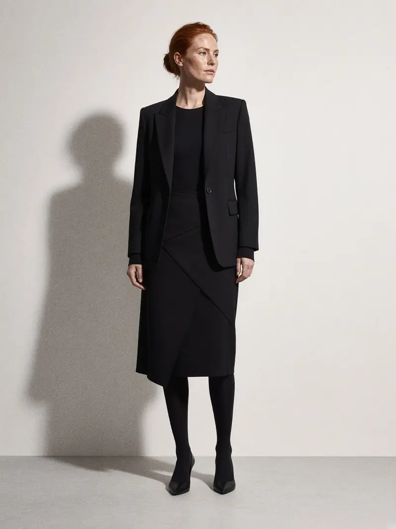 Illustration, mannequin fictif : Femme en blazer noir ajusté, body noir, jupe portefeuille noire au genou, collants opaques et escarpins pointus noirs.
