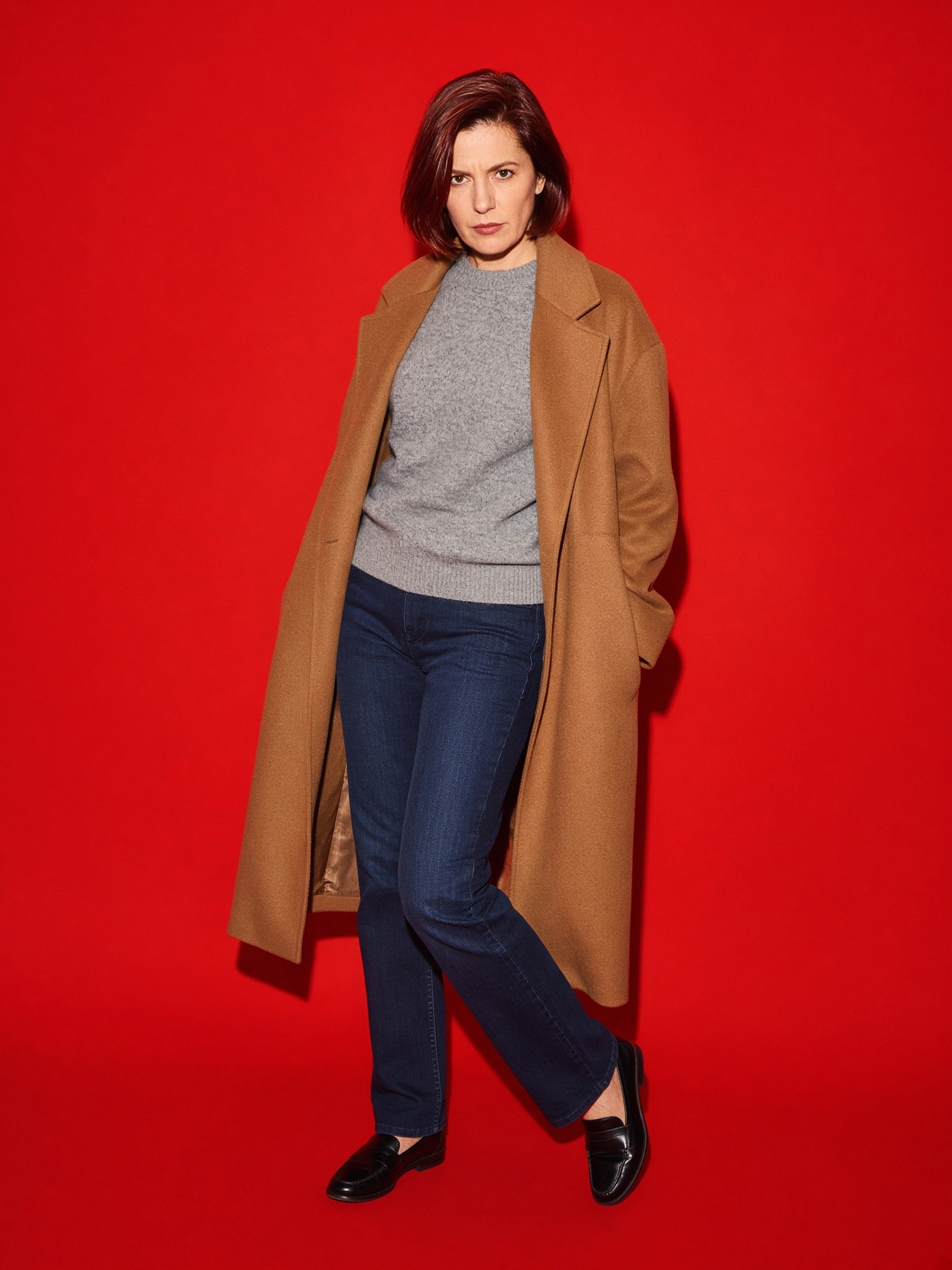 Mannequin en manteau de laine camel, pull gris et jean droit indigo, debout dans une chambre d'hôtel au mur beige.