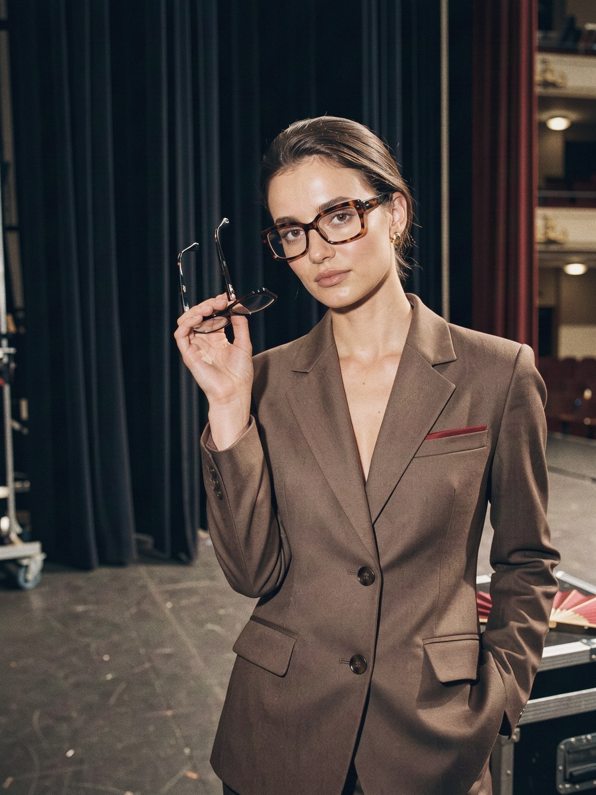 Mannequin au visage rond comparant deux montures de lunettes devant un tailleur marron taupe