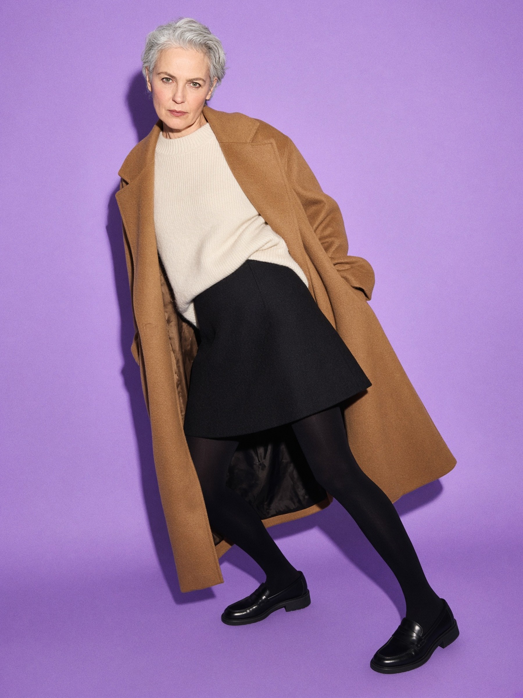 Mannequin en mini jupe de laine noire, pull écru ample, collants opaques et long manteau camel dans un couloir d'hôtel