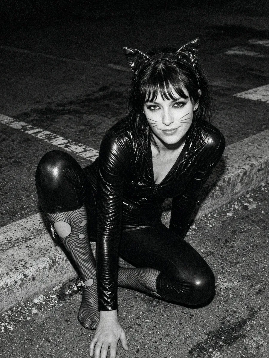 Catwoman accroupie Déguisement Catwoman sexy, combinaison noire brillante, oreilles de chat et résille filée, accroupie sur un trottoir