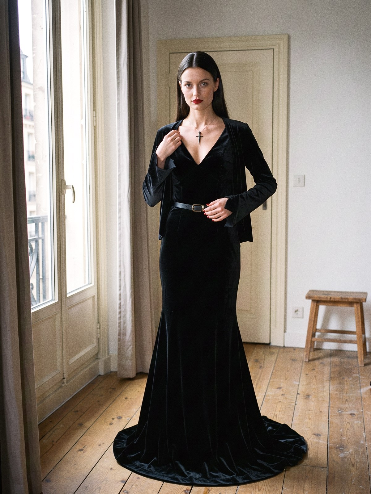 Mannequin de mode ajustant un long cardigan noir sur une robe noire à col en V et manches pointues, maquillage pâle et bouche sombre.