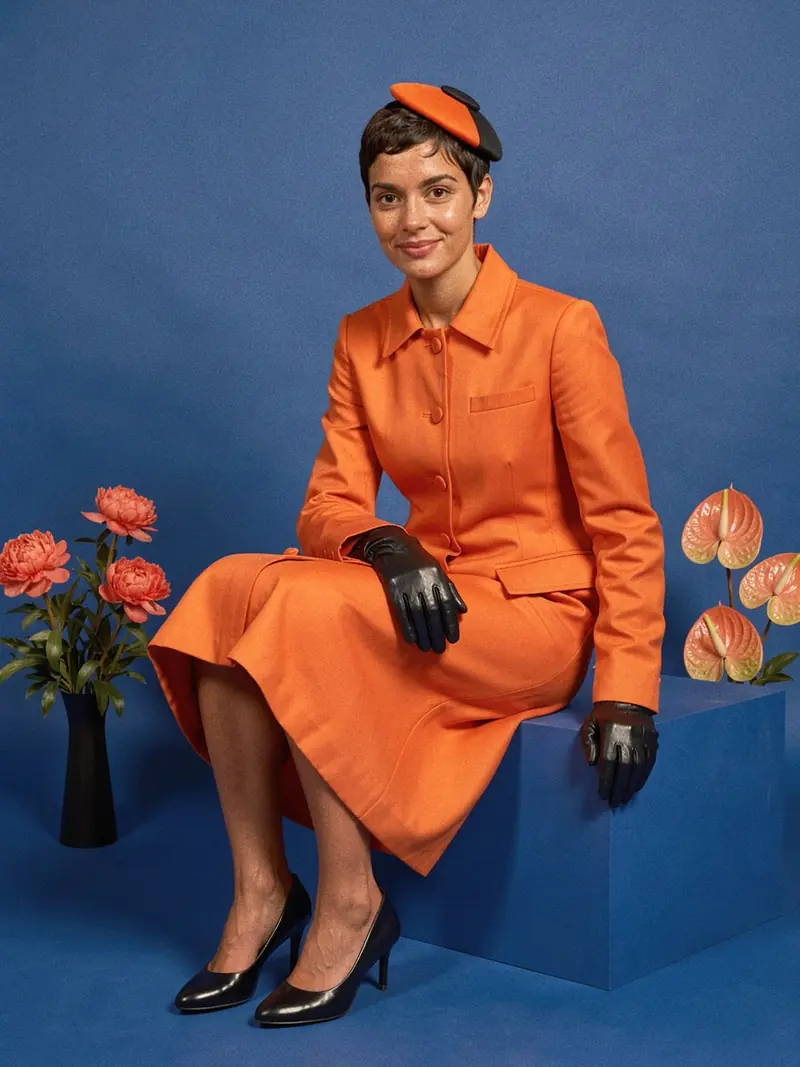 Illustration, mannequin fictif : Femme en manteau-robe orange vif boutonné jusqu'au genou et jupe midi assortie, petit bibi plat orange et noir, gants noirs et escarpins noirs.