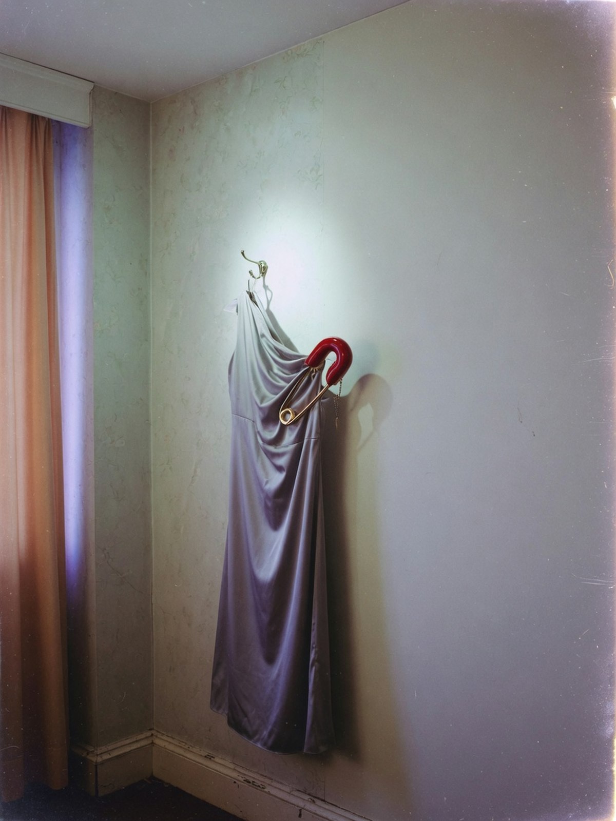 Robe satinée fermée par une grande épingle décorative, suspendue seule sur un mur nu, éclairée au flash direct.