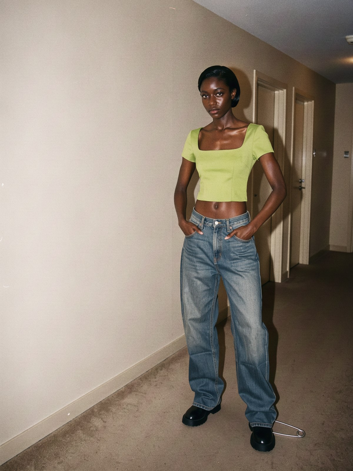 Mannequin portant un jean baggy taille basse avec un haut court structuré et une chaussure épaisse, dans un couloir nu éclairé au flash.