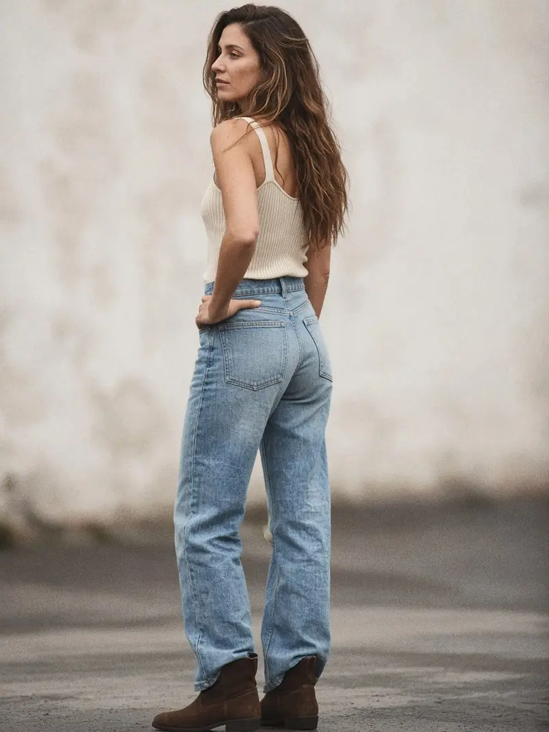 Illustration, mannequin fictif : Femme debout de trois quarts dos en jean bootcut délavé clair et débardeur côtelé écru, la main glissée à plat dans la ceinture arrière.