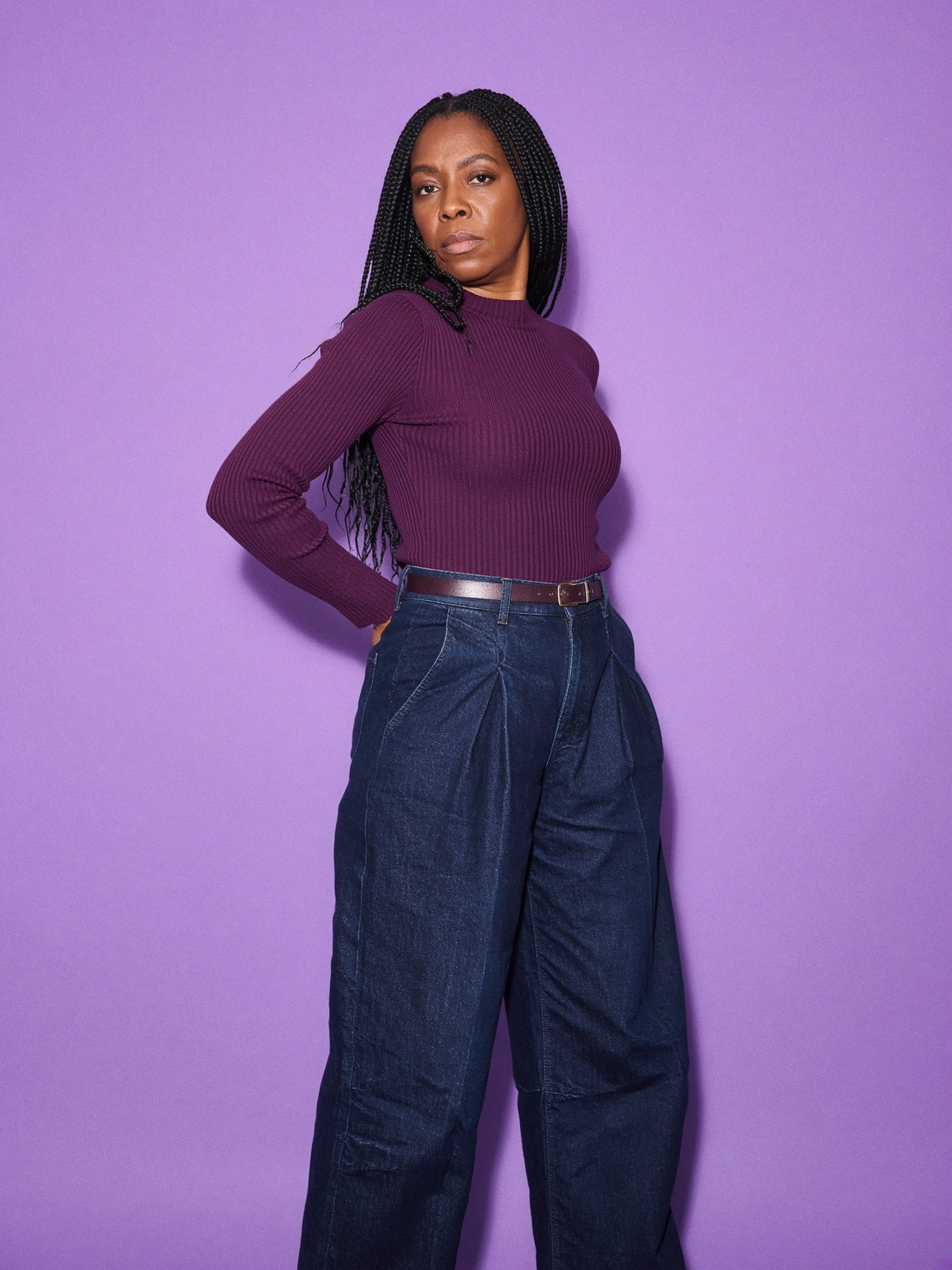 Mannequin debout dans une chambre d'hôtel nue, jean wide leg indigo taille haute à pinces, haut côtelé ajusté rentré, ceinture fine.