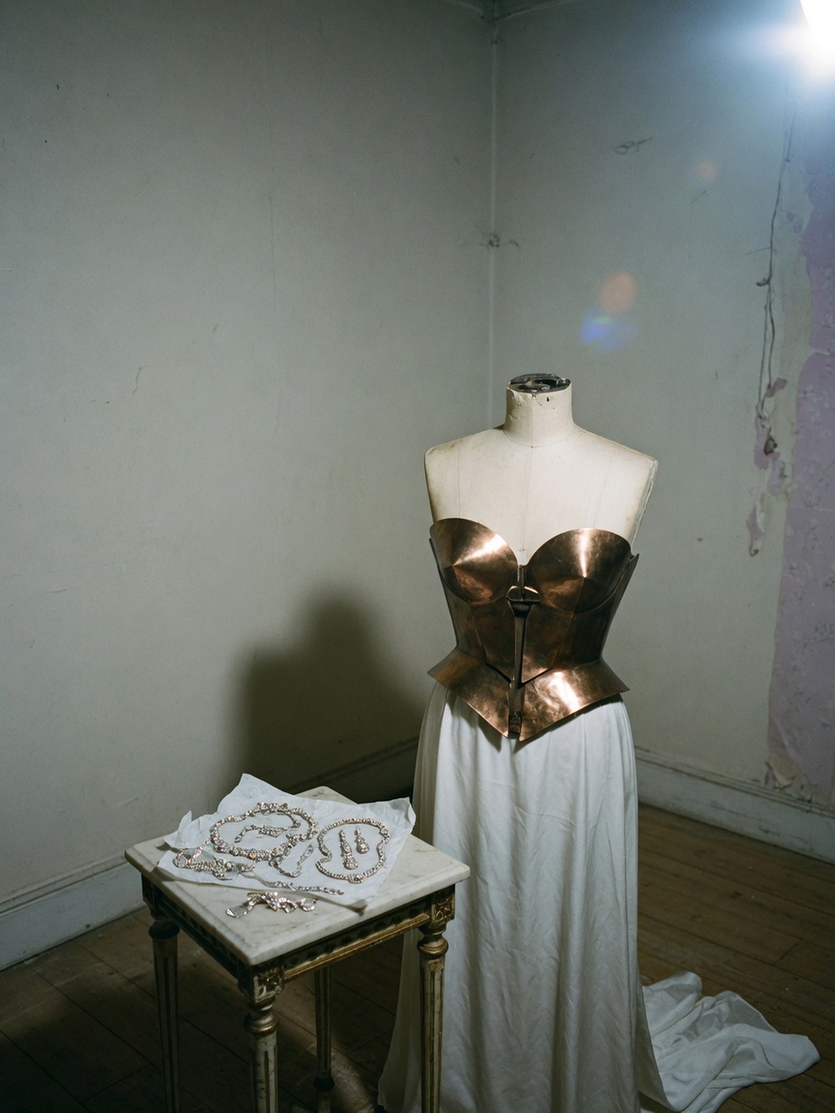 Nature morte évoquant le bustier bronze et la jupe blanche du look Met Gala 2026 de Léna Situations, présentés sur un buste de couturier avec des bijoux posés à côté.