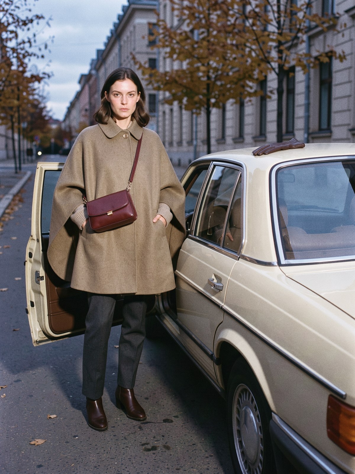 Mannequin portant un manteau cape en laine kaki avec un sac porté en bandoulière devant, près d'une voiture au crépuscule.