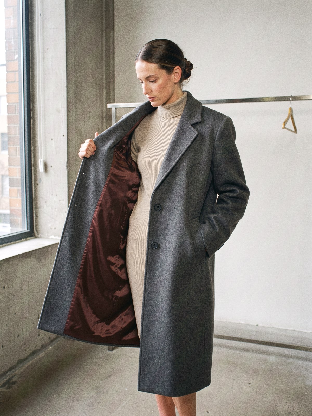 Mannequin tenant un pan de manteau en laine charcoal vers la lumière, doublure oxblood visible