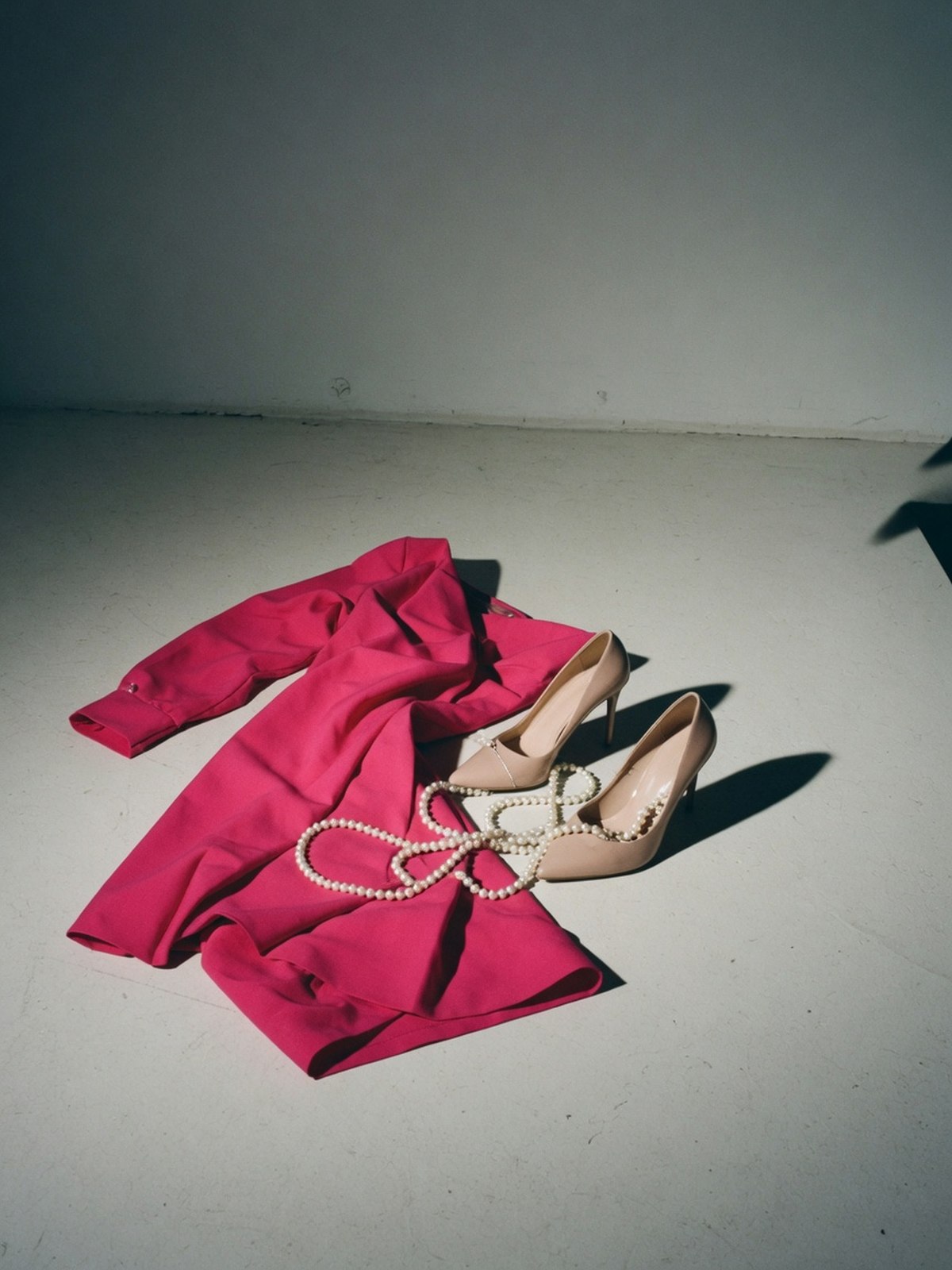 Détail éditorial d'un pan de robe fuchsia, d'escarpins nude à talons aiguilles et d'un collier de perles disposés au sol.