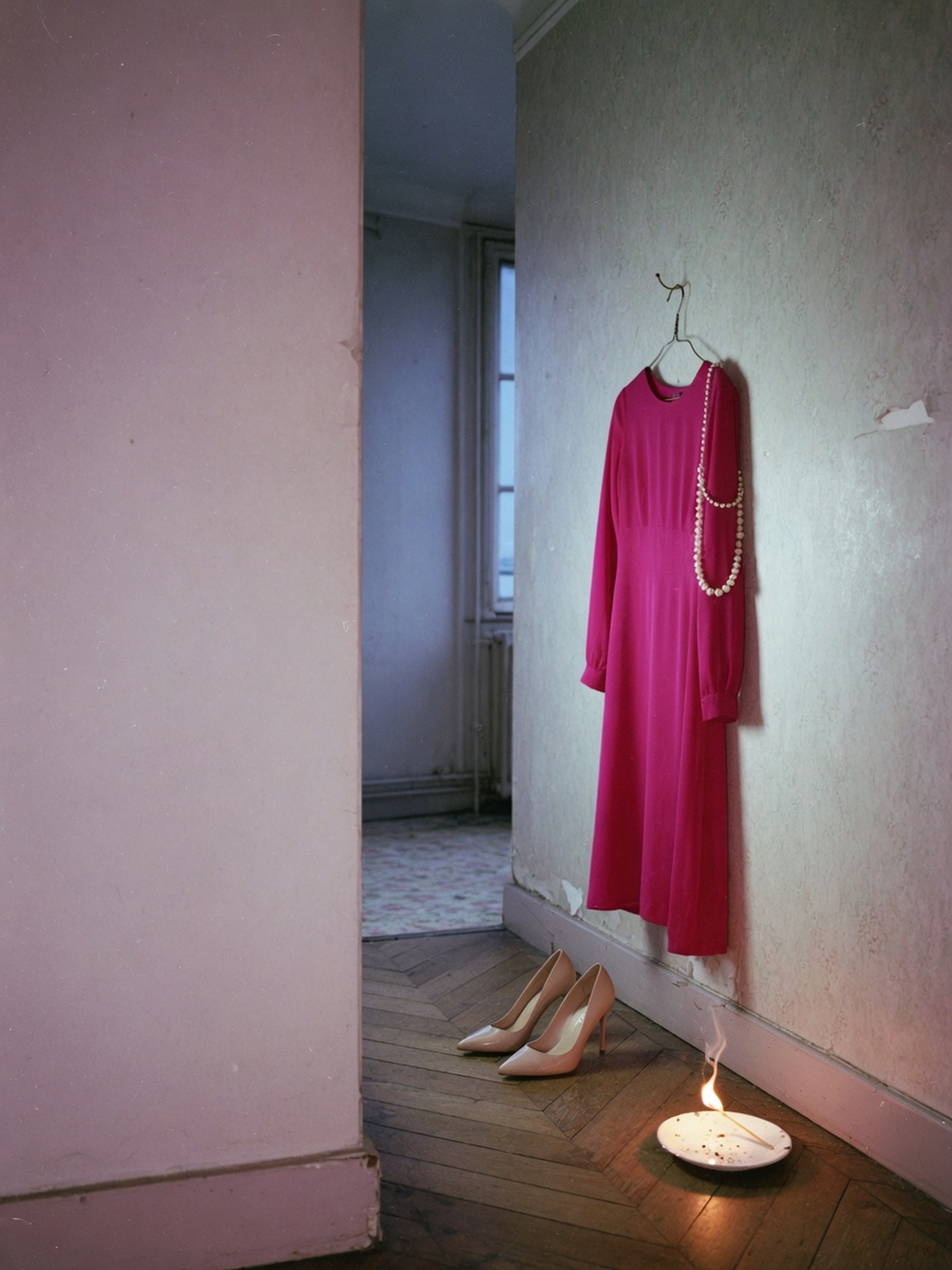 Nature morte éditoriale d'une robe longue fuchsia, d'escarpins nude à talons aiguilles et d'un collier de perles, dans un couloir d'appartement parisien à la lumière de flash.