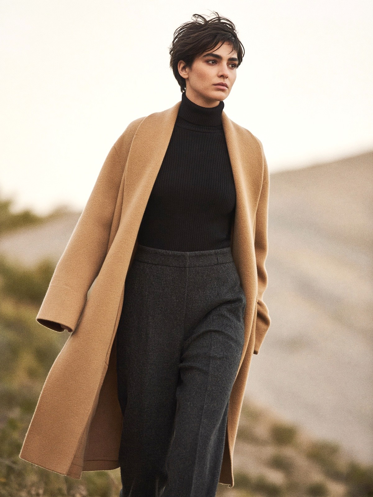 Mannequin en manteau-cardigan camel ouvert sur un col montant noir et un pantalon droit gris, mains dans les poches, les pans croisés à la taille.