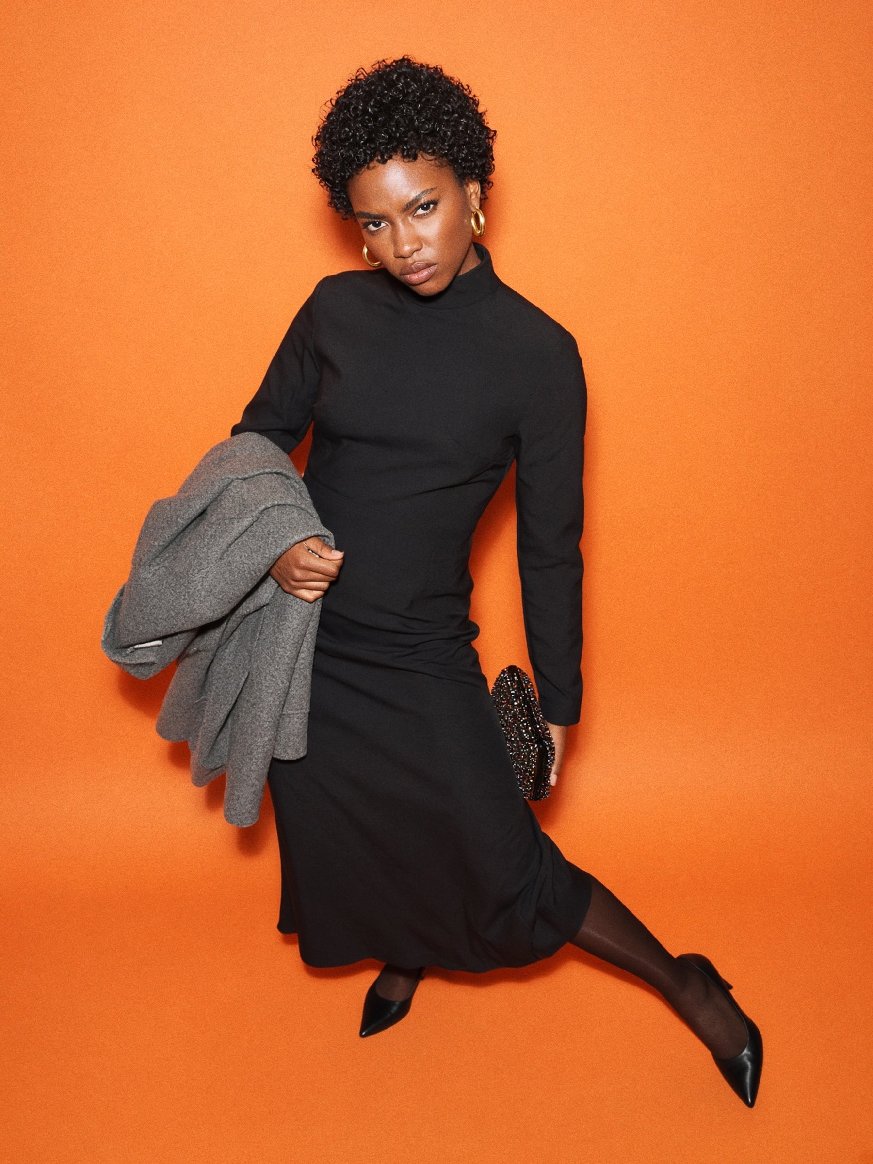 Mannequin en robe midi noire en crêpe, collants foncés et escarpins fermés, manteau plié sur le bras, dans un couloir d'hôtel au crépuscule.