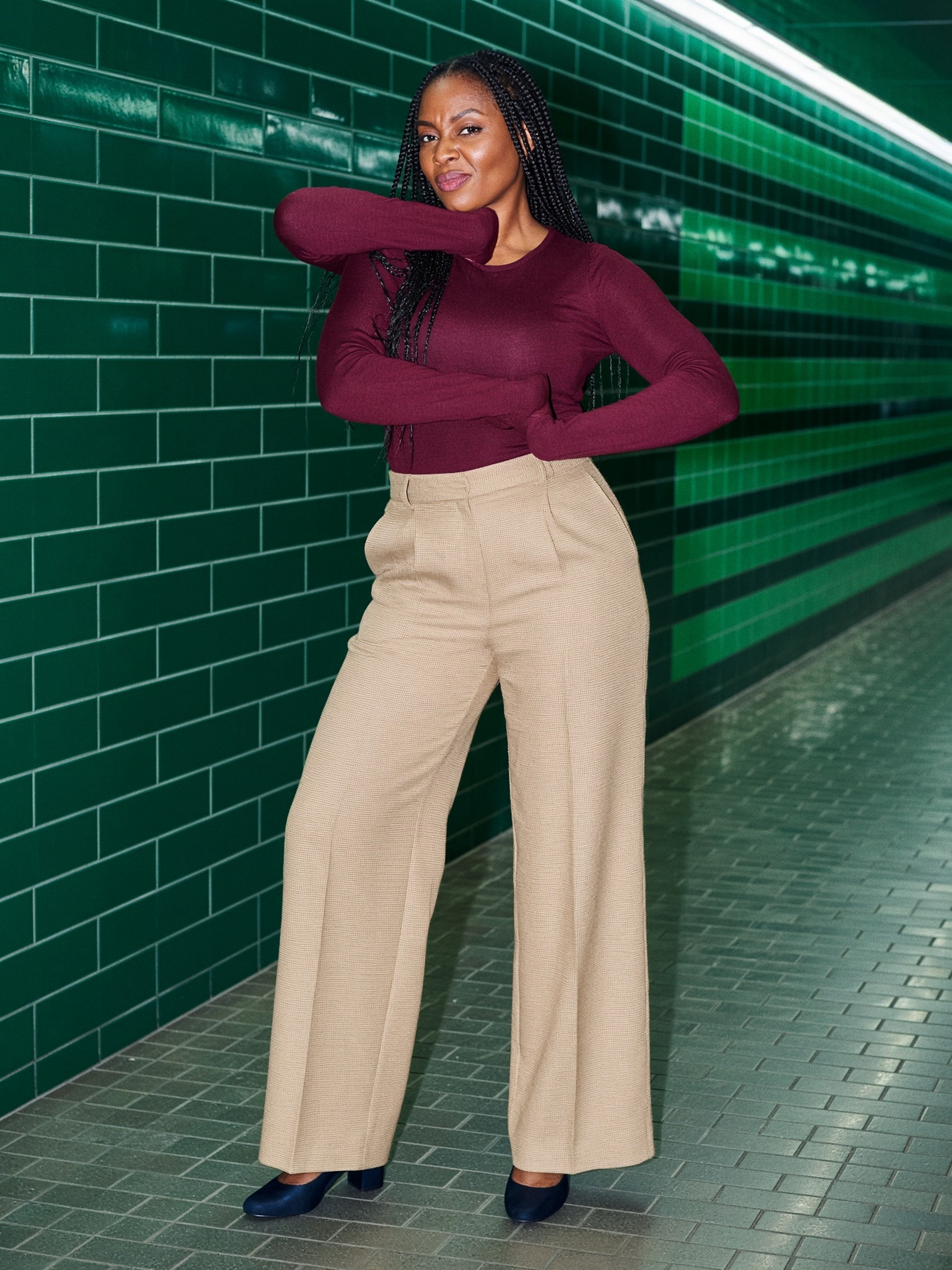 Mannequin en pantalon beige large taille haute et maille bordeaux, debout dans un couloir d'hôtel, visage entièrement visible.