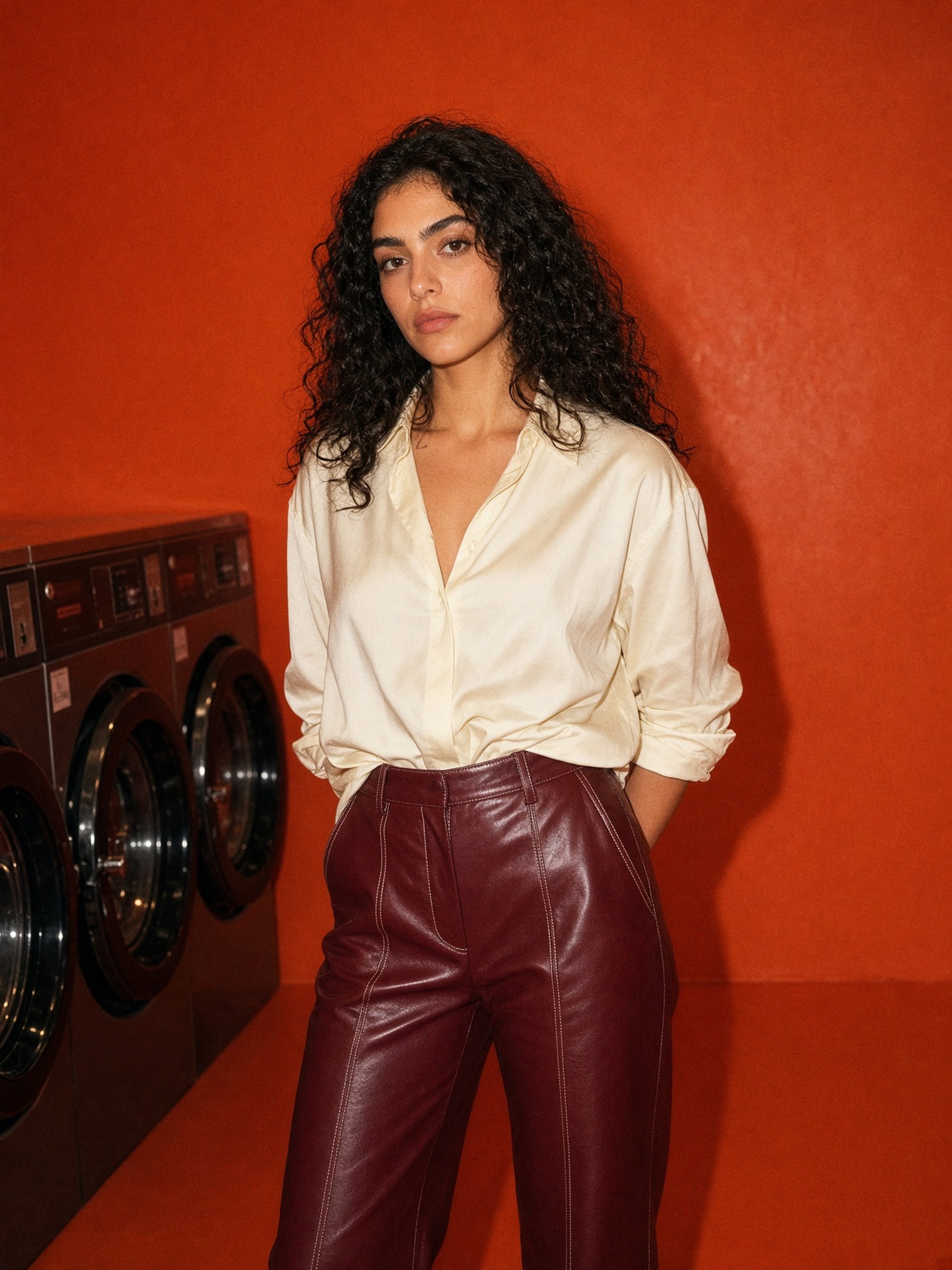 Mannequin en pantalon en cuir bordeaux taille haute et chemise en soie crème, debout dans une chambre d'hôtel au crépuscule