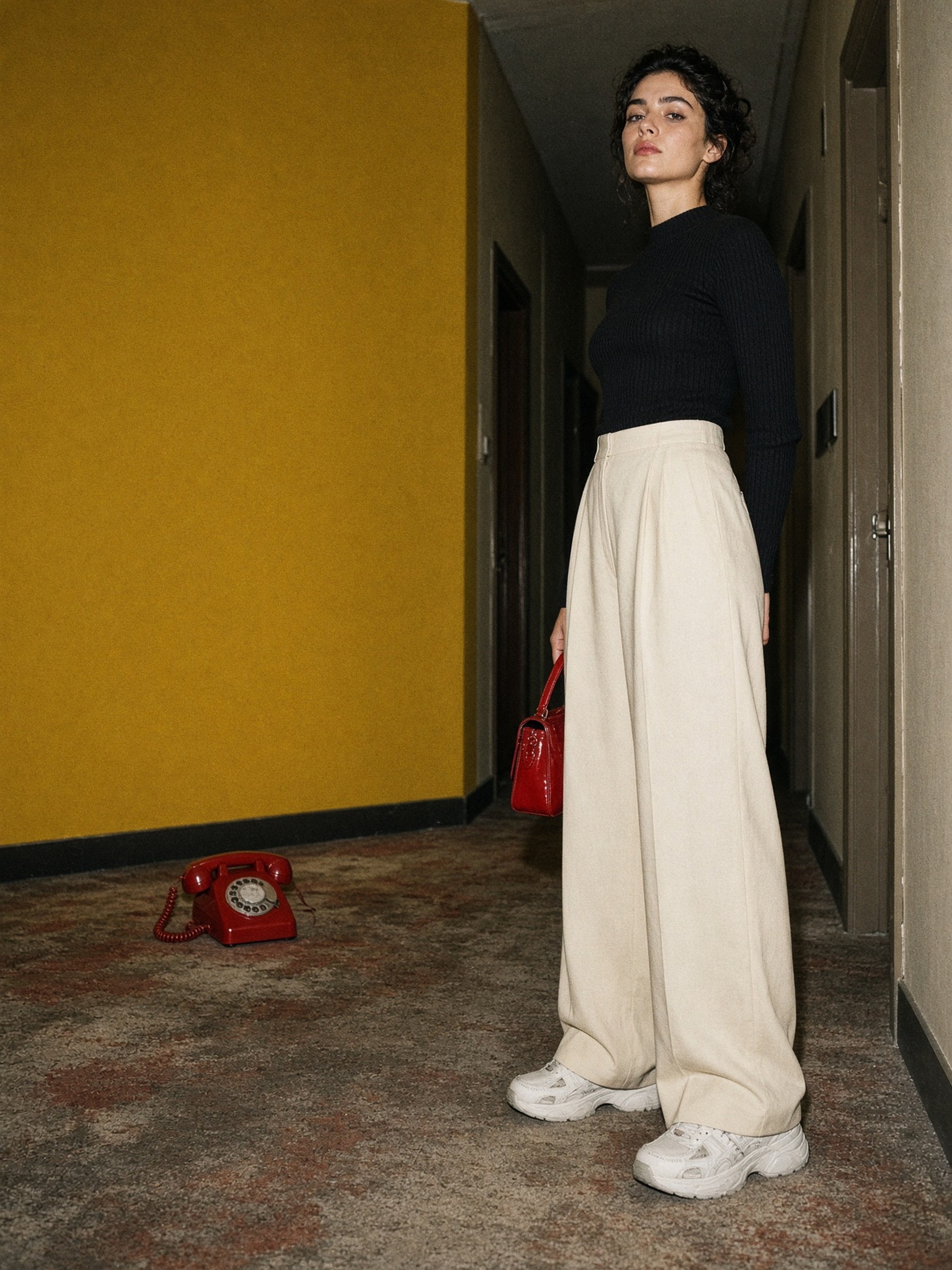 Mannequin en pantalon large écru fluide, haut ajusté noir et baskets blanches basses, debout dans un couloir d'hôtel.