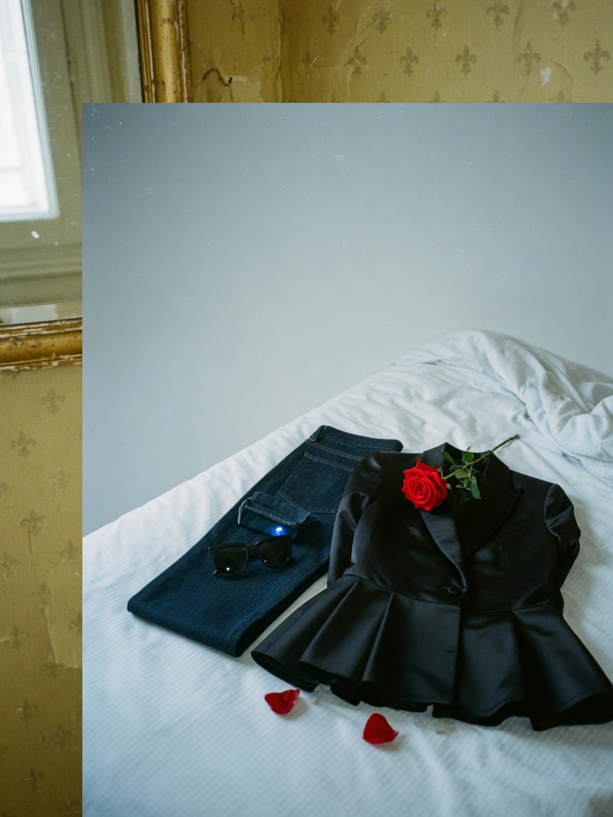 Veste péplum noire structurée, jean droit et lunettes de soleil disposés sur un lit d'hôtel, dans une mise en scène épurée