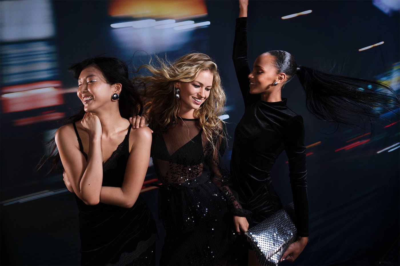 Trois femmes en tenues de fête noires, velours, tulle transparent et sequins, dansant dans la ville la nuit, campagne Only