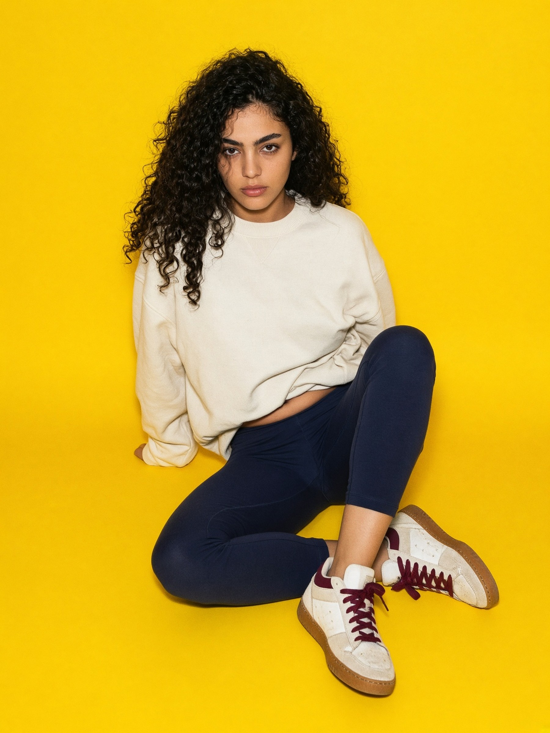 Mannequin en legging bleu marine et sweat écru oversize, baskets en cuir blanc cassé à détails bordeaux, dans un studio nu éclairé au flash.