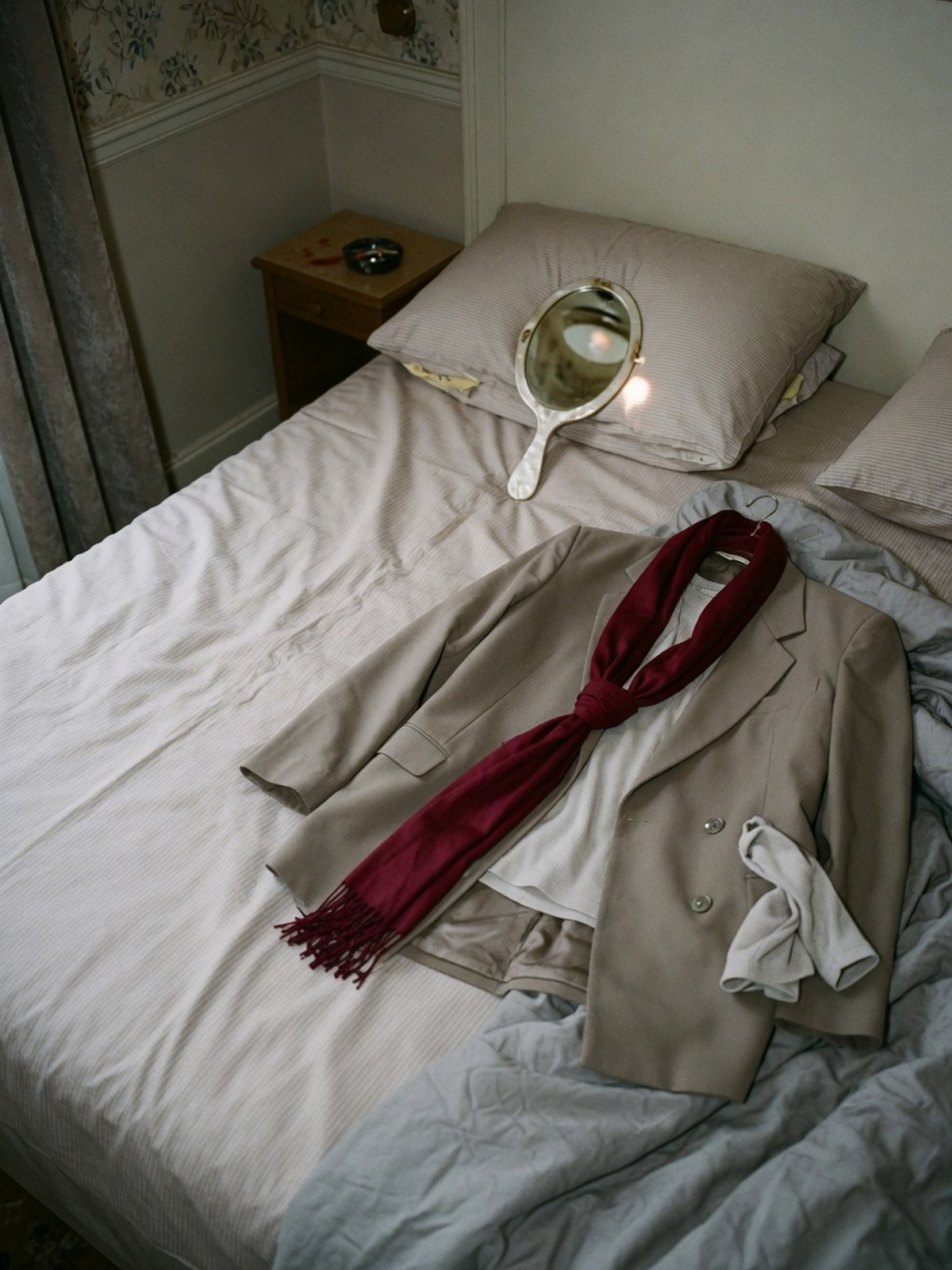 Un blazer oversize superposé à une base légère, posé sur un lit défait, avec un foulard bordeaux comme touche de couleur.