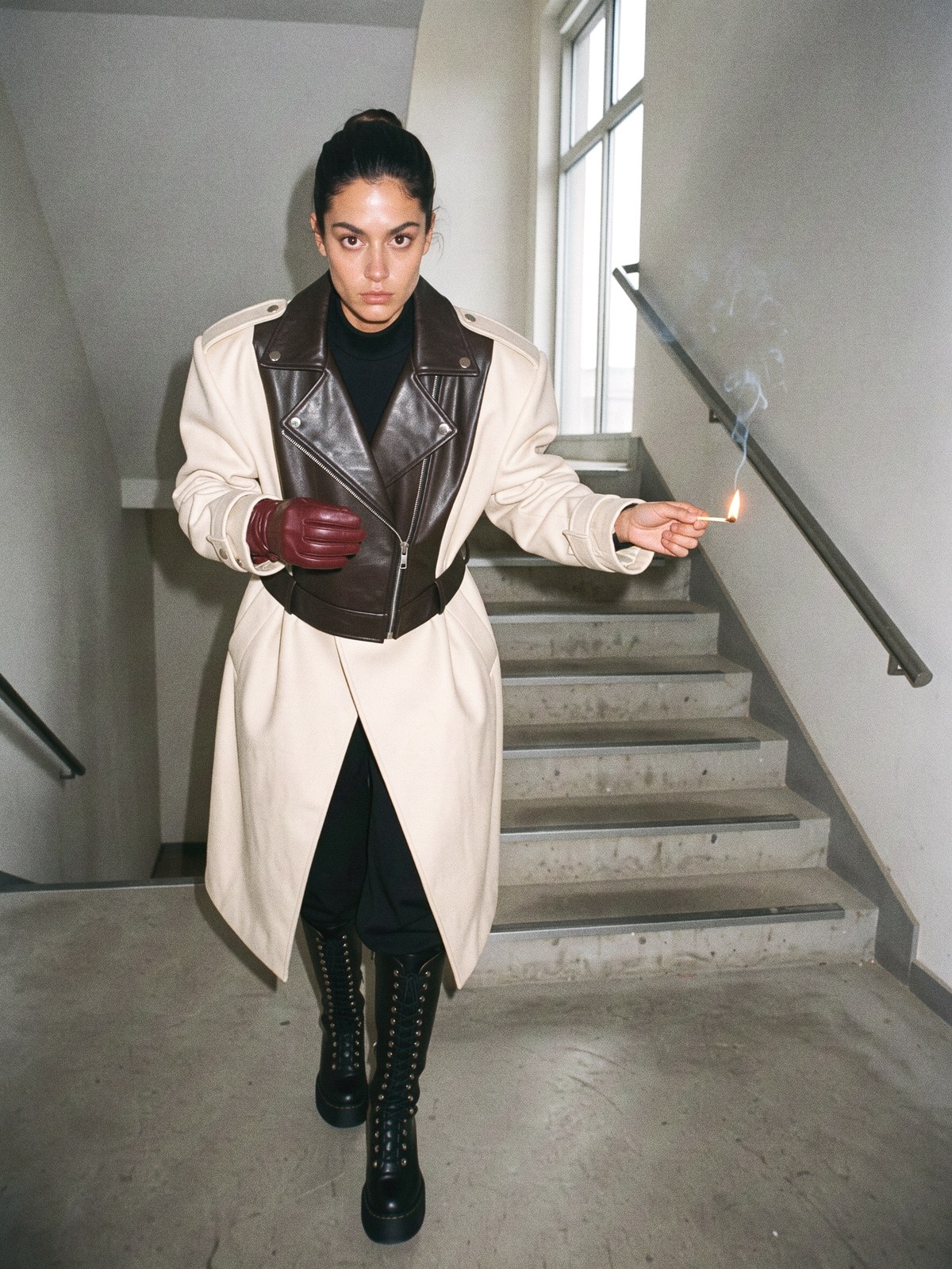 Mannequin en manteau crème oversize et pantalon noir large, un gant de cuir bordeaux tenu contre la poitrine, posture éditoriale dans un escalier en béton.