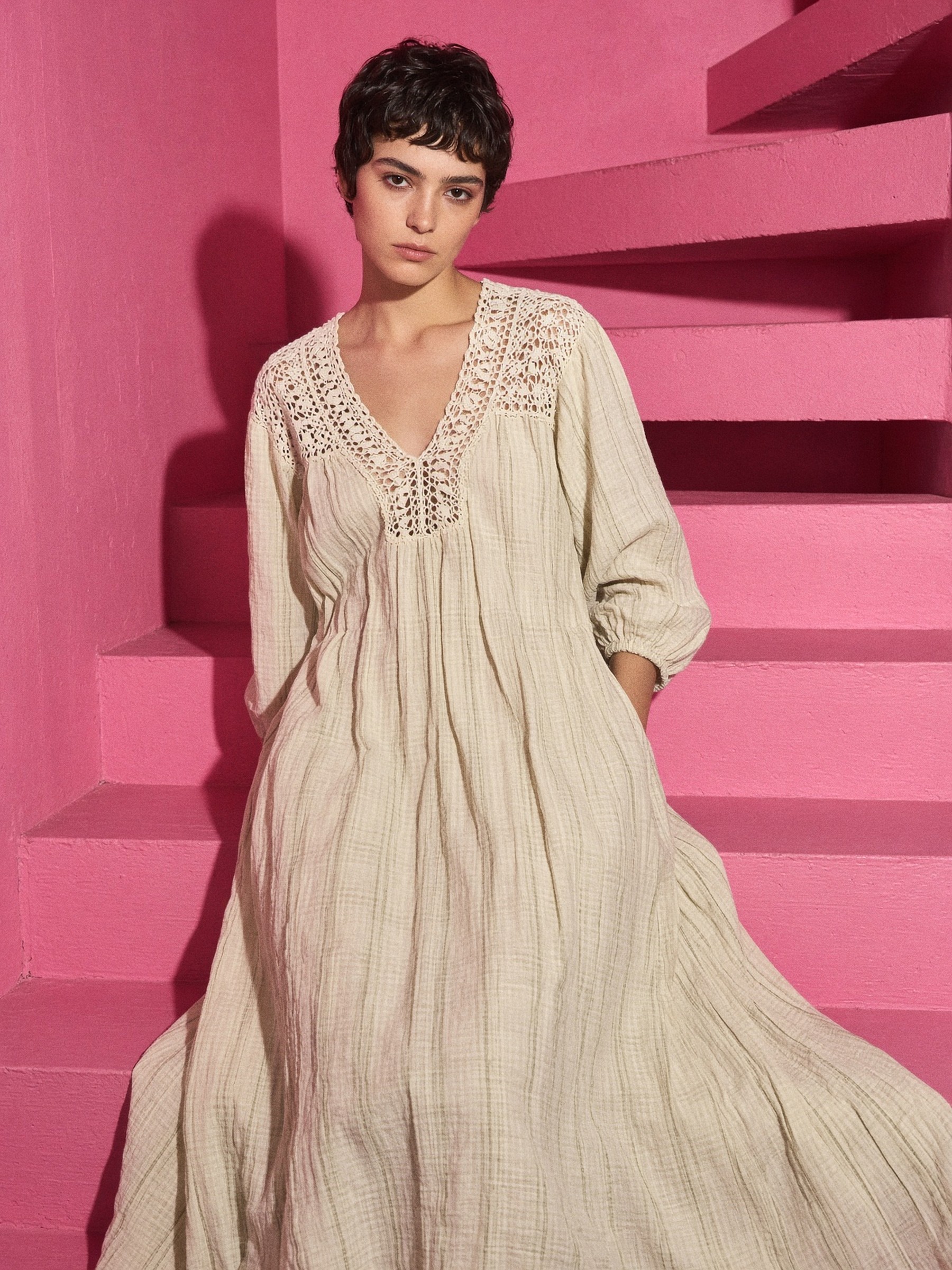 Mannequin en robe longue bohème en coton gaze crème à empiècement de dentelle, debout dans une chambre d'hôtel, un fauteuil bordeaux en arrière-plan
