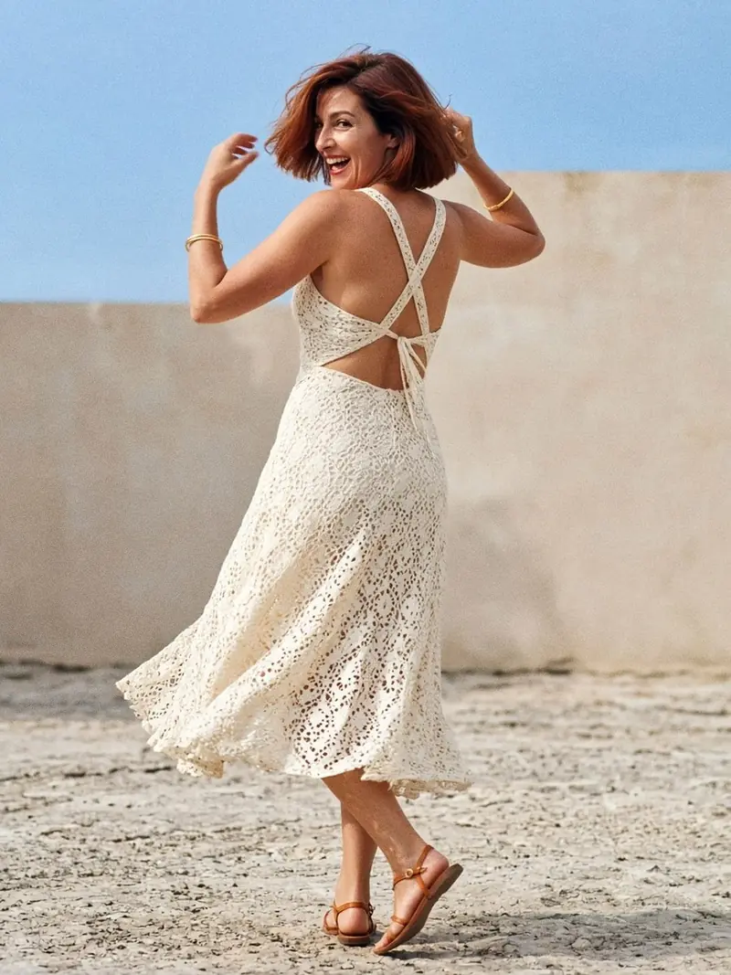 Illustration, mannequin fictif : Femme en mouvement, bras levés, en robe midi en dentelle de coton ivoire doublée, bretelles croisées sur un dos ouvert jusqu'à la taille, sandales plates.