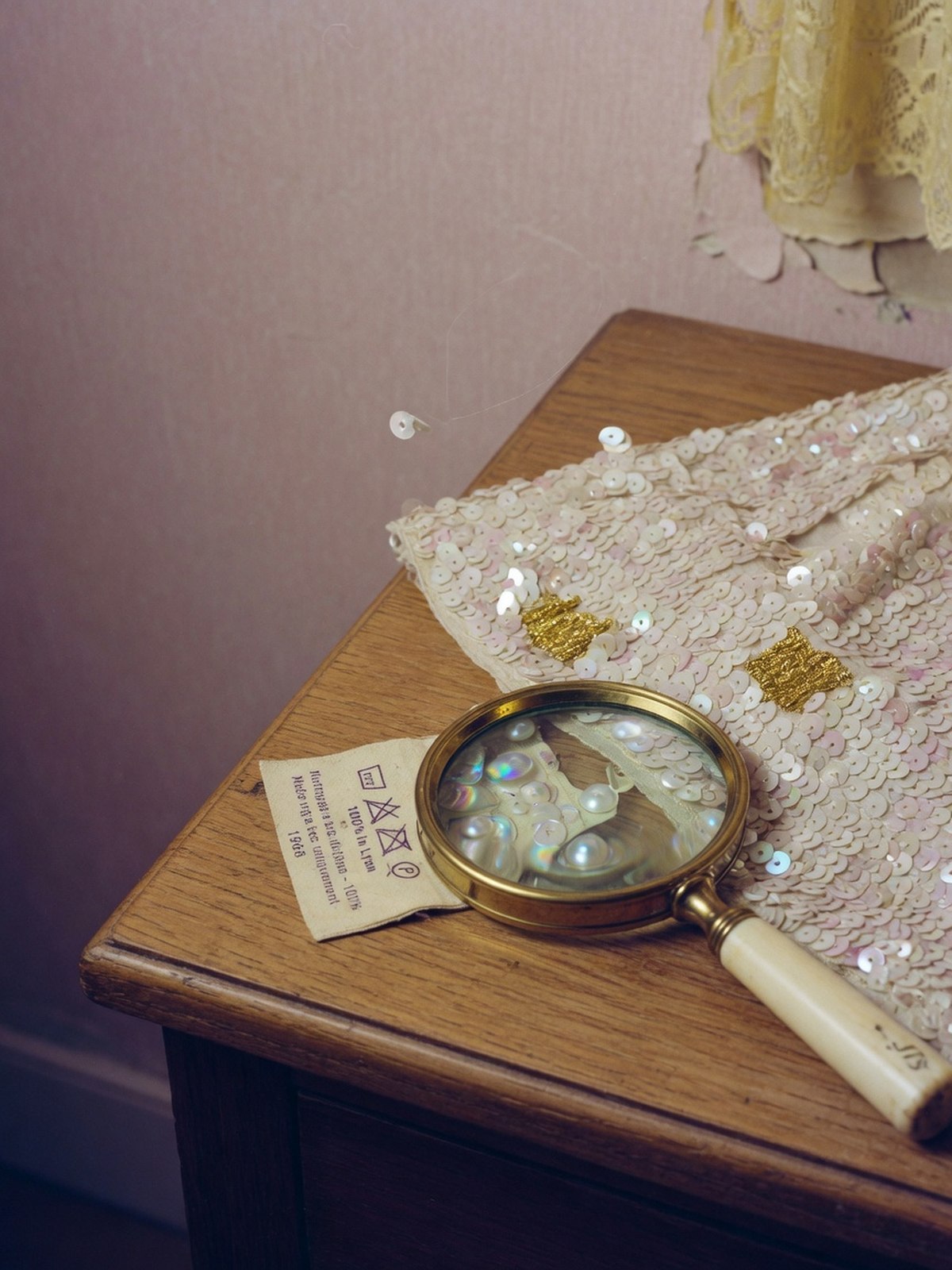 Détail d'un ourlet de robe vintage à paillettes nacrées, étiquette de composition et loupe posées à côté sur une coiffeuse en bois.