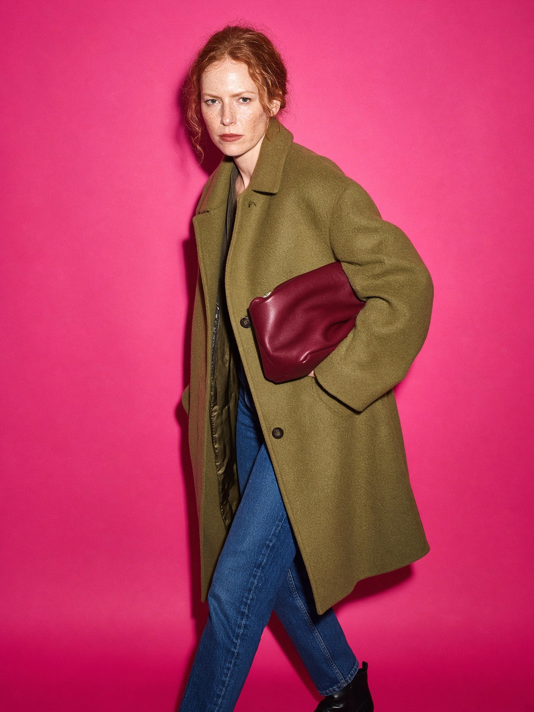 Mannequin de profil dans un couloir, manteau marron chocolat et mini sac baguette bordeaux porté sous le bras.