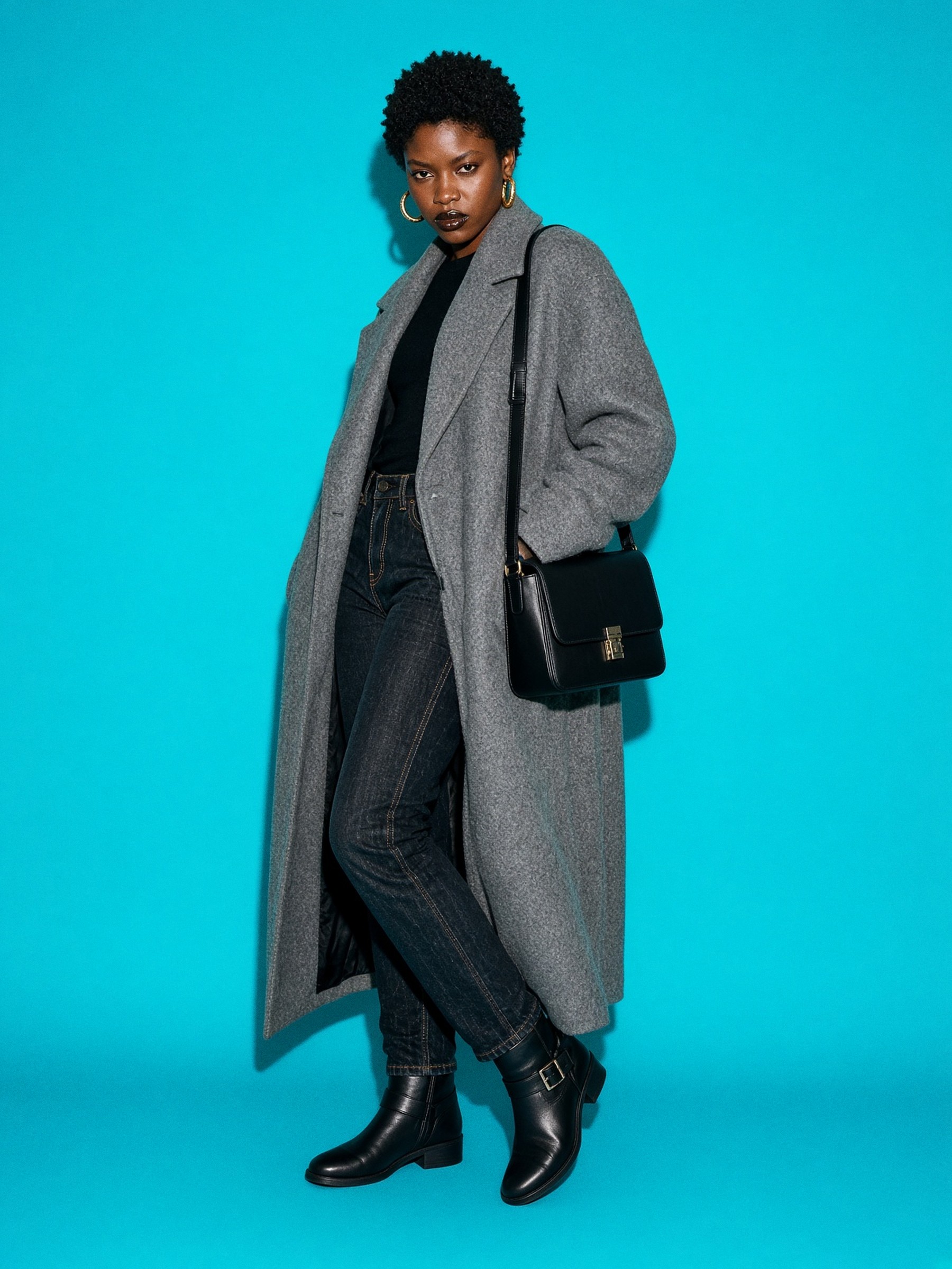 Mannequin de face dans un couloir nu, en manteau long de laine grise porté ouvert sur un jean brut et des bottines noires.