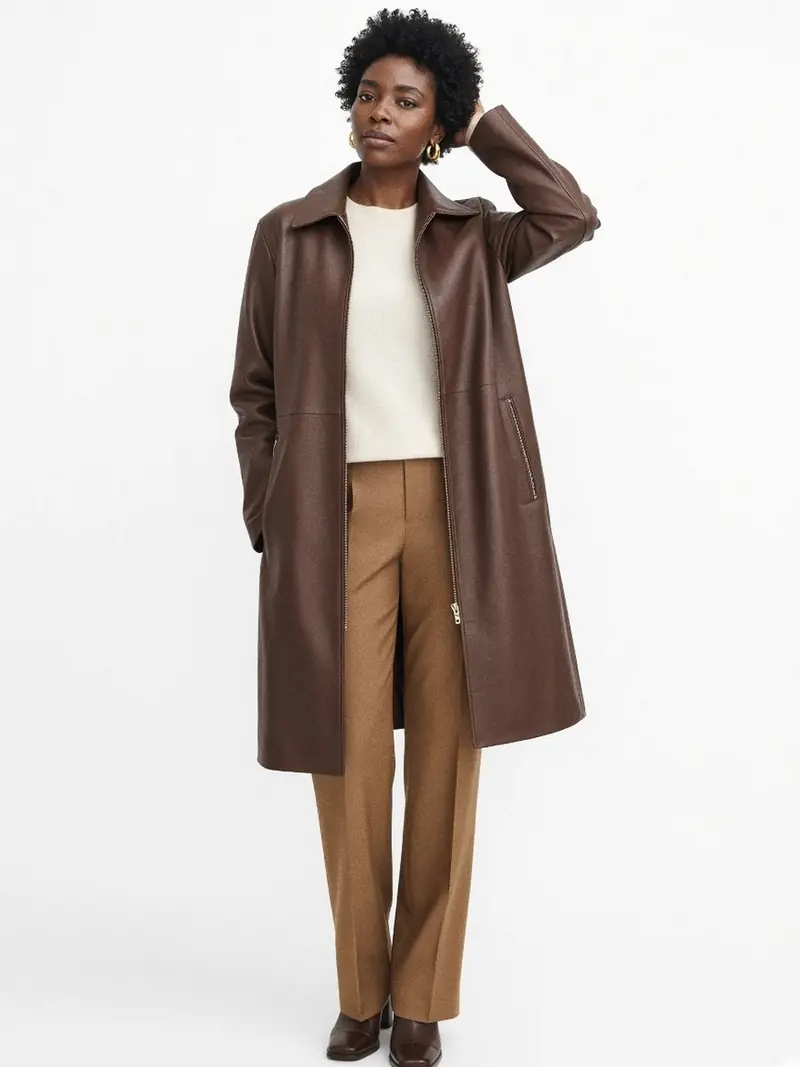 Illustration, mannequin fictif : Femme en veste en cuir marron chocolat longueur genou, fermeture centrée et poches zippées, un bras levé, pull ivoire et pantalon camel