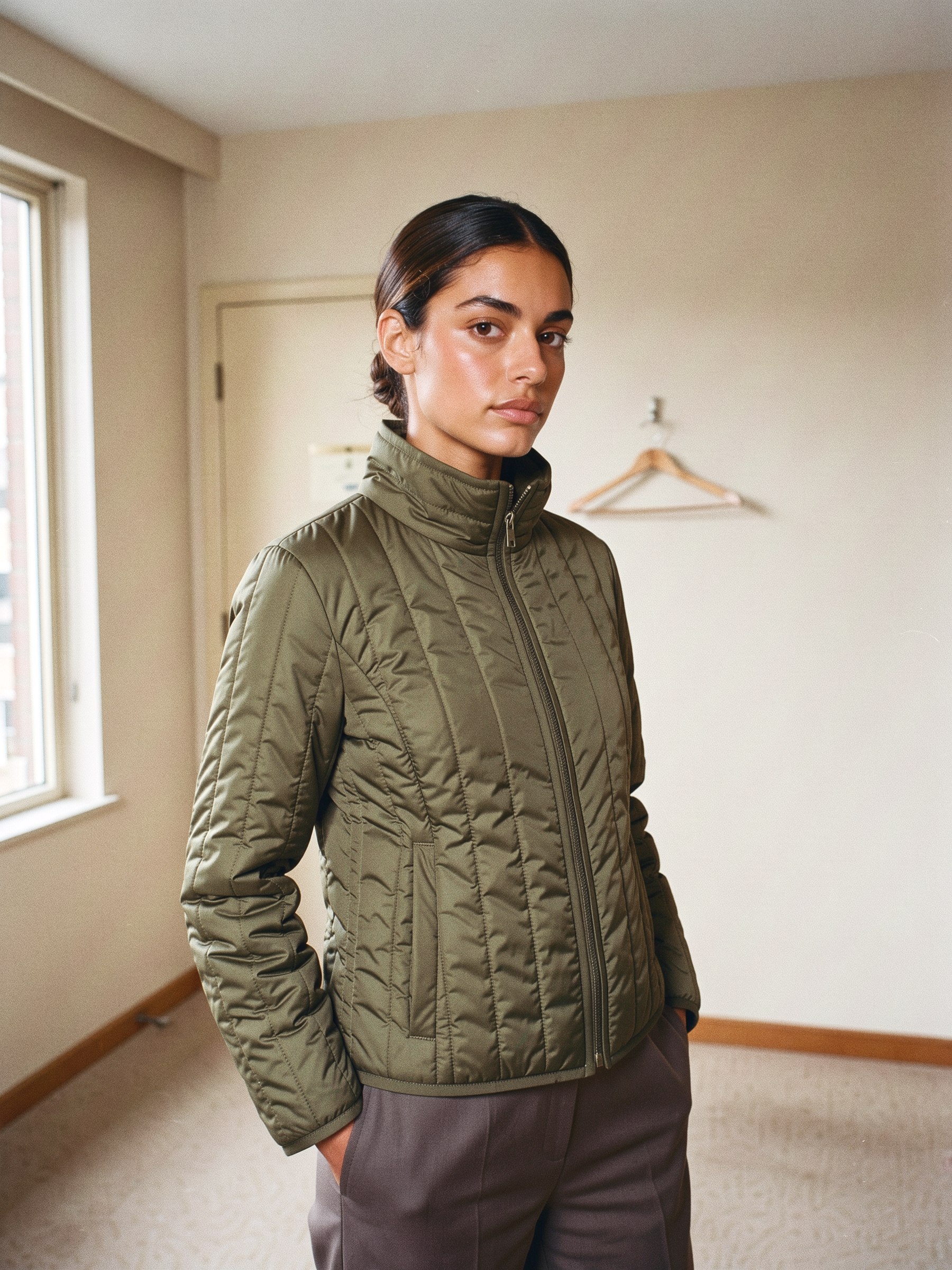 Mannequin en veste matelassée courte vert olive à col montant, debout dans une chambre d'hôtel aux murs beiges
