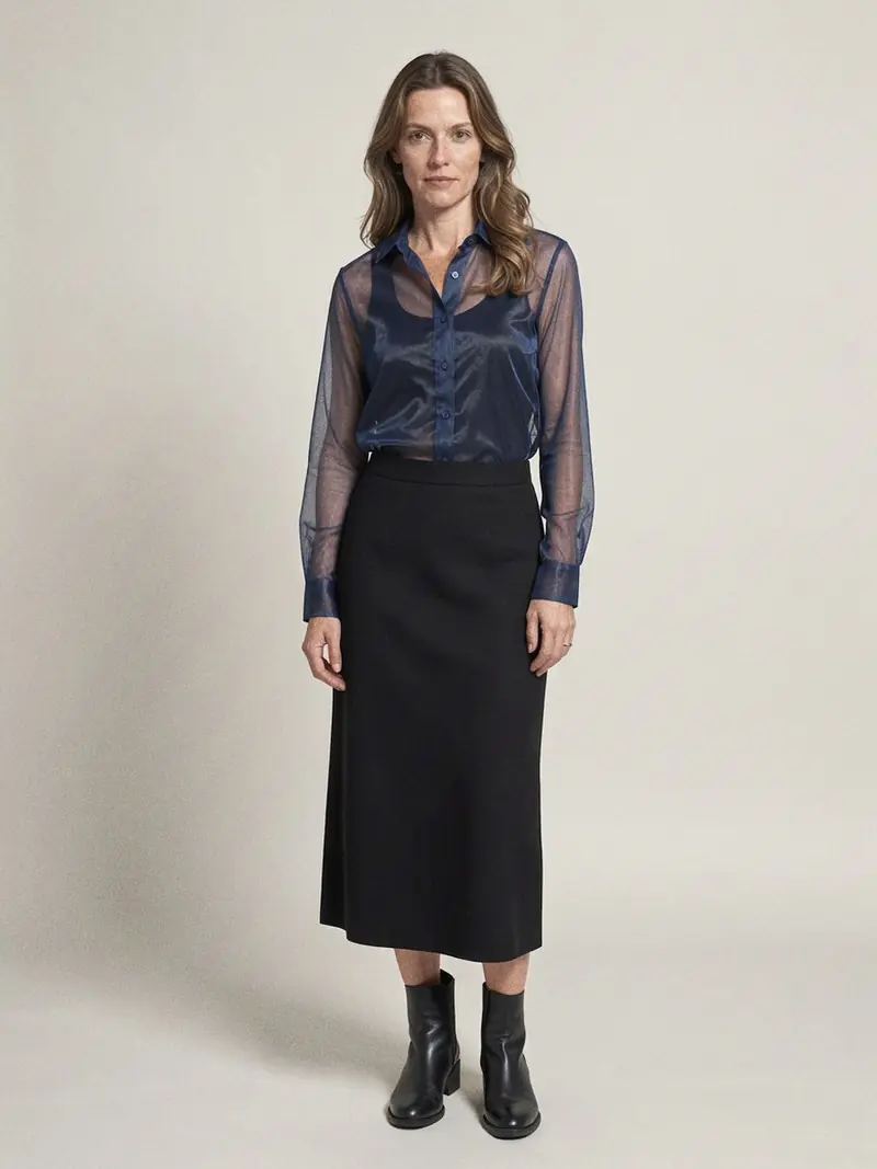 Illustration, mannequin fictif : Femme de trois quarts en chemise en organza bleu nuit transparente boutonnée sur un haut en maille noire mate, jupe droite noire et bottines noires.