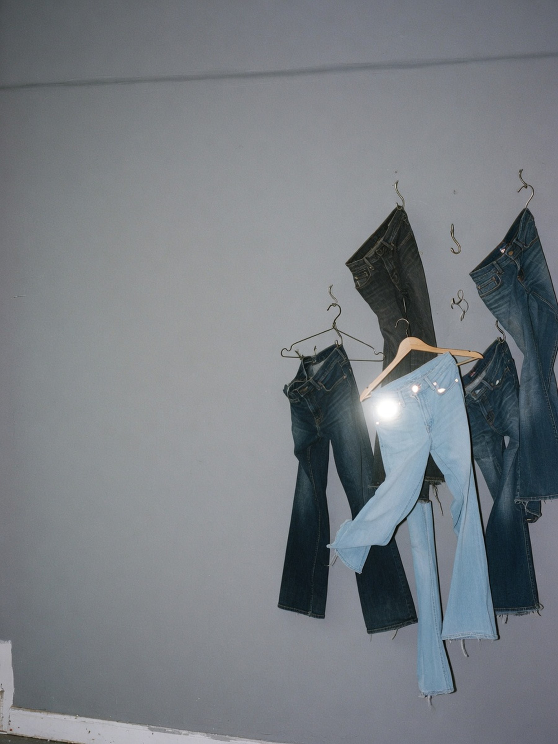 Nature morte éditoriale de plusieurs jeans bootcut bleus suspendus à des crochets métalliques dans un studio nu, un des jeans éclairé par un flash direct qui accentue sa teinte bleue face aux autres plus sourds.